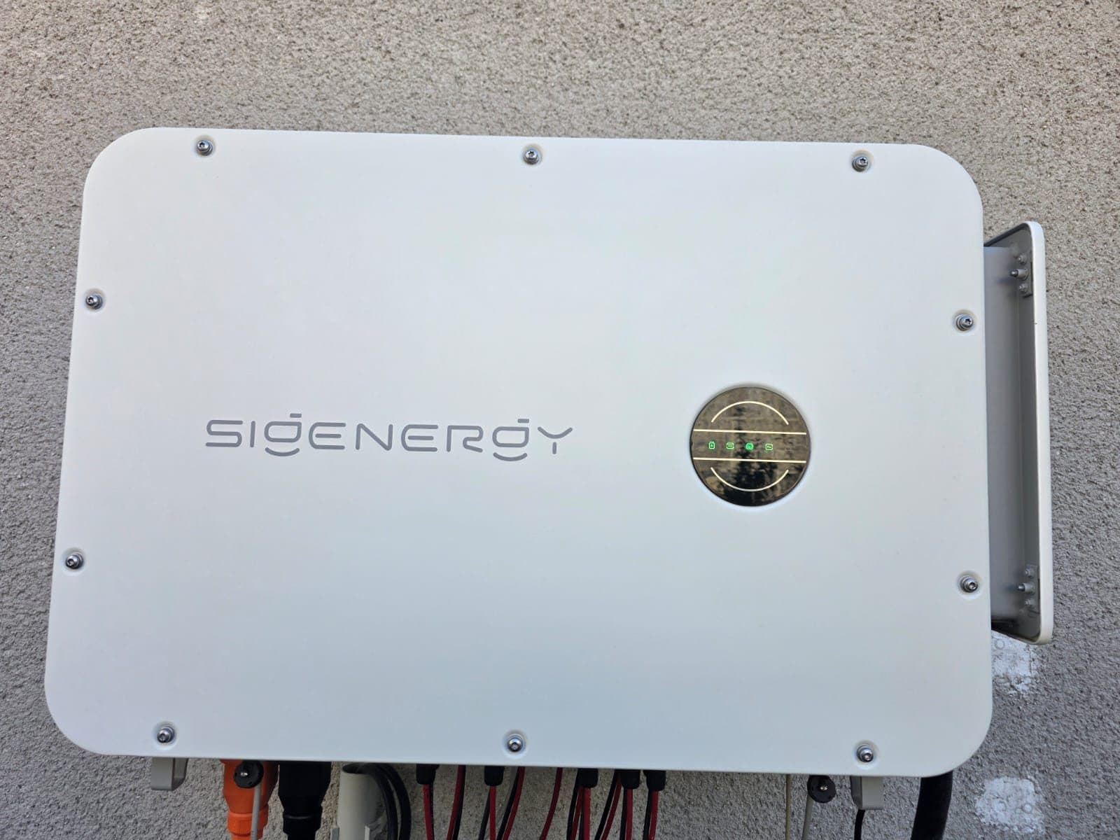 Weißer Sigenergy-Wechselrichter frontal an einer Putzwand, die runde Anzeige leuchtet grün, an der Unterseite stecken rote und schwarze Solarleitungen