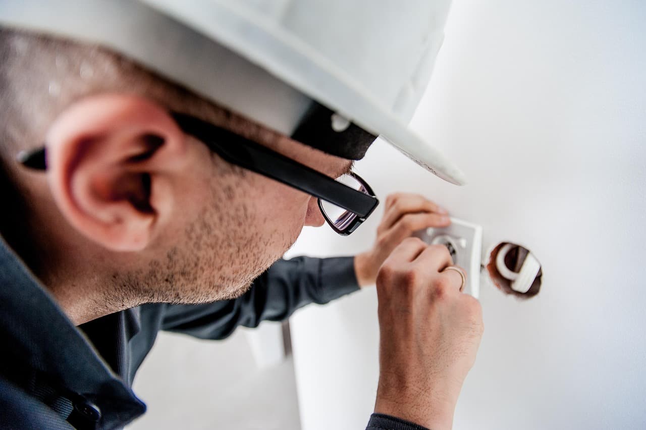 Nahaufnahme eines Elektrikers mit weißem Helm und Brille, der eine Steckdose in eine weiße Wand einsetzt